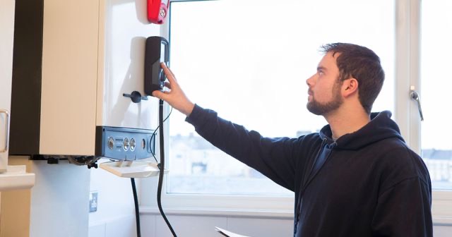 Een monteur bedient het paneel van een cv-ketel aan de muur, met een raam op de achtergrond